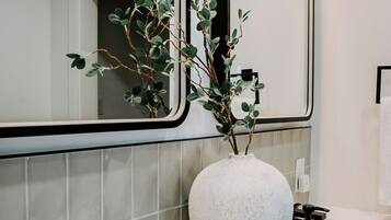 A modern bathroom with a white sink, black faucets, a large mirror, and a white vase with green foliage.