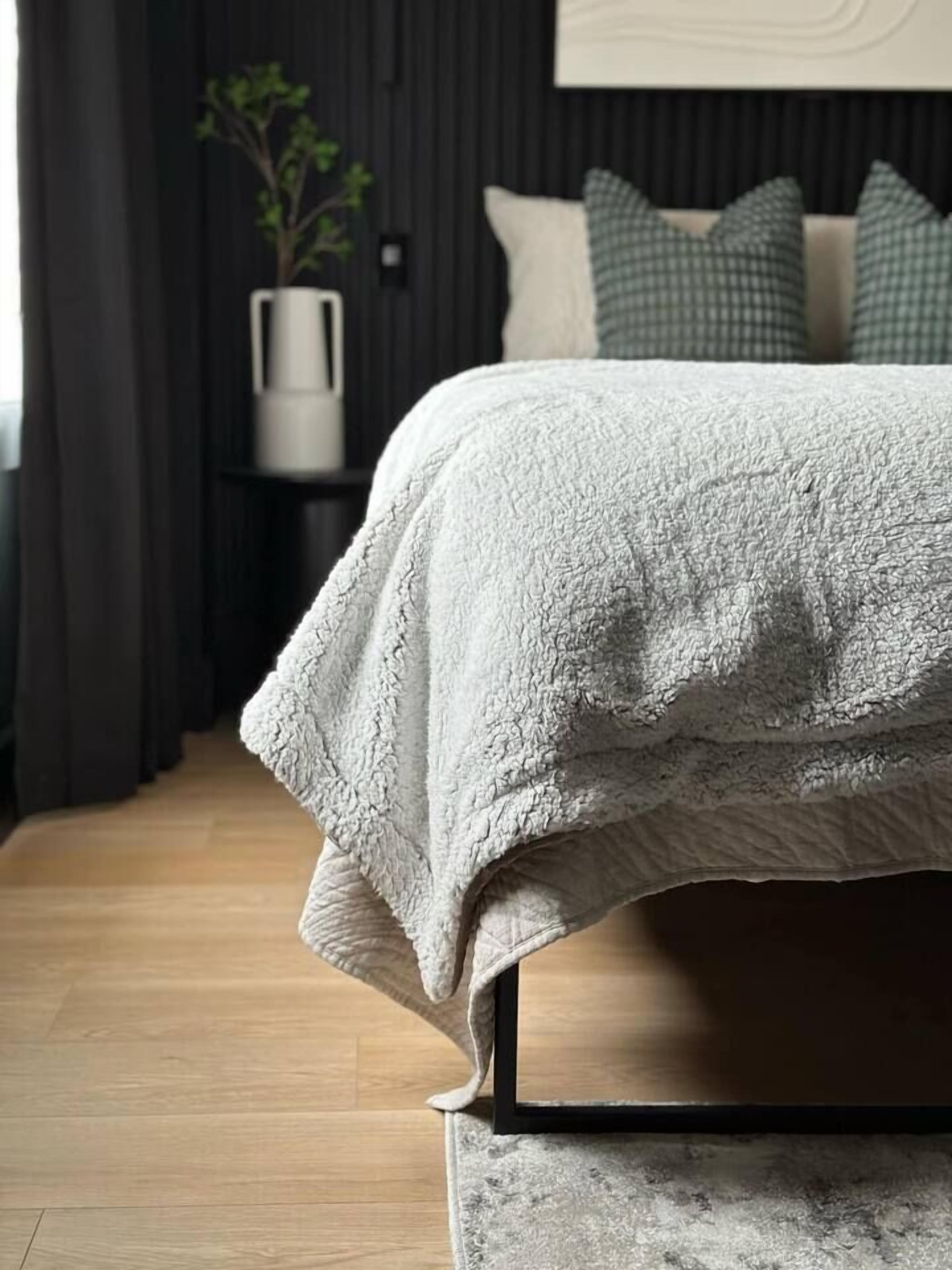 Una cama con una manta blanca con textura, una pared oscura y un suelo de madera.