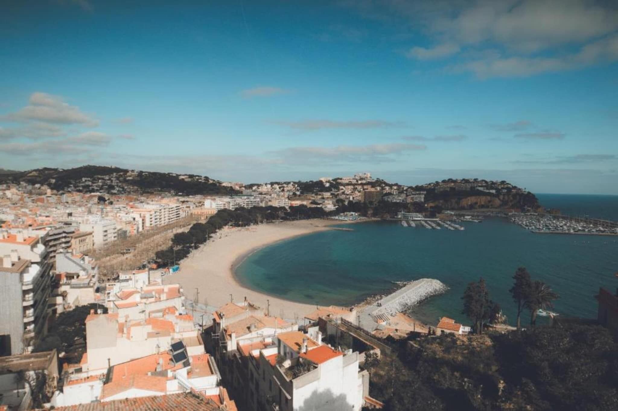 Paisatge urbà costaner amb platja, edificis i un turó amb construccions.