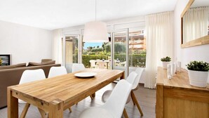 Un salon moderne avec une table à manger en bois, des chaises blanches, un canapé et une grande fenêtre donnant sur un jardin et des immeubles.