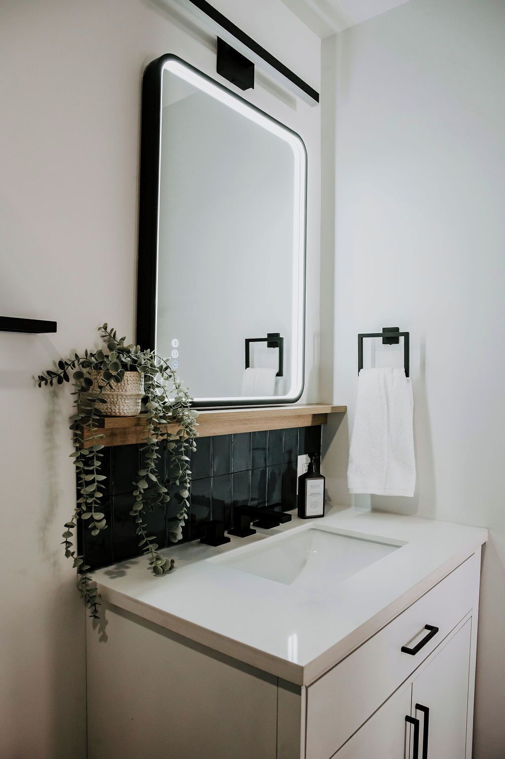 Une salle de bain moderne avec un grand miroir, un lavabo blanc et une étagère en bois sur laquelle est posée une plante.