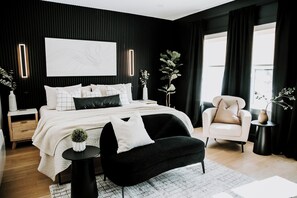 A modern bedroom with a black and white color scheme, featuring a large bed, a black bench, a white armchair, and a bedside table with a vase.