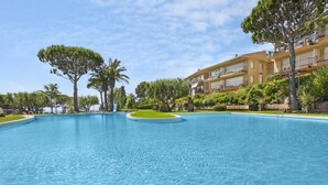 Une grande piscine aux eaux claires, avec un îlot circulaire au centre, entourée d’une végétation luxuriante et bien entretenue, et bordée d’un immeuble à plusieurs étages doté de balcons.