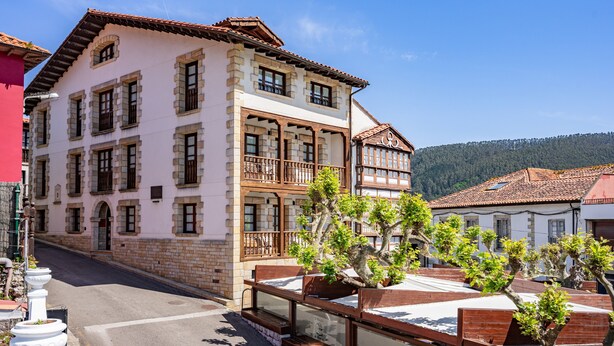 A multi-story building with balconies, a street in front, and a mountainous backdrop.