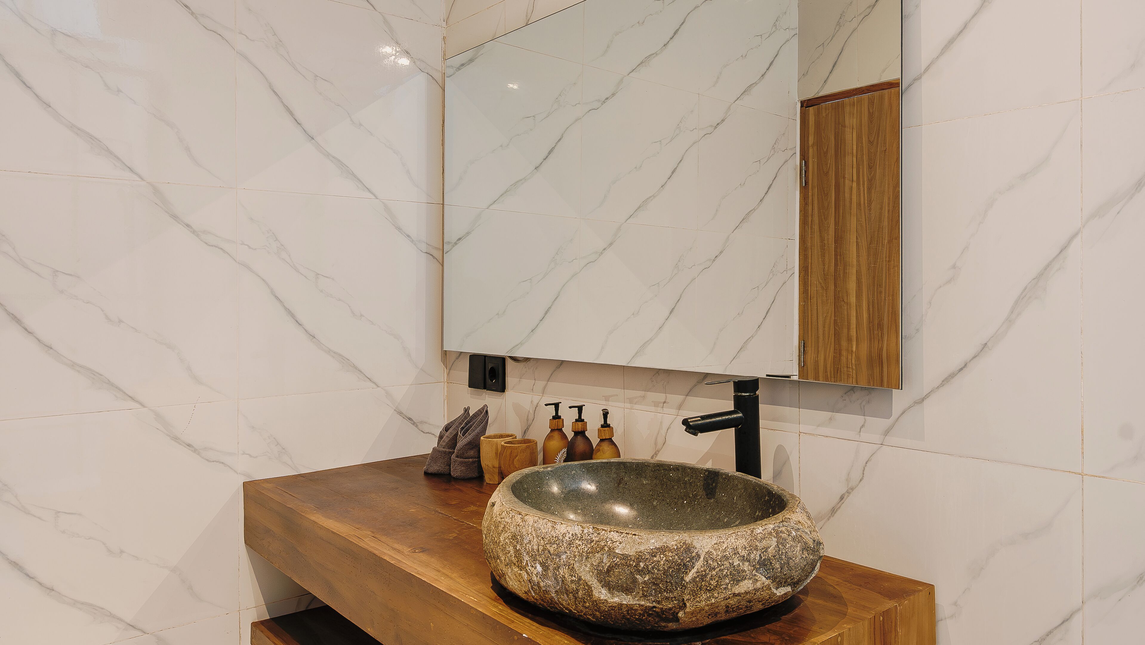 A bathroom with a marble wall, a wooden vanity, a stone basin, and a wooden cabinet above the mirror.