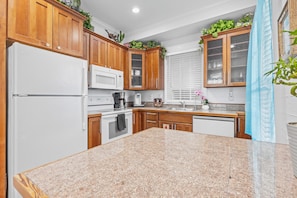A kitchen with wooden cabinets, a granite countertop, a white refrigerator, a microwave, a coffee maker, and a window with blinds.