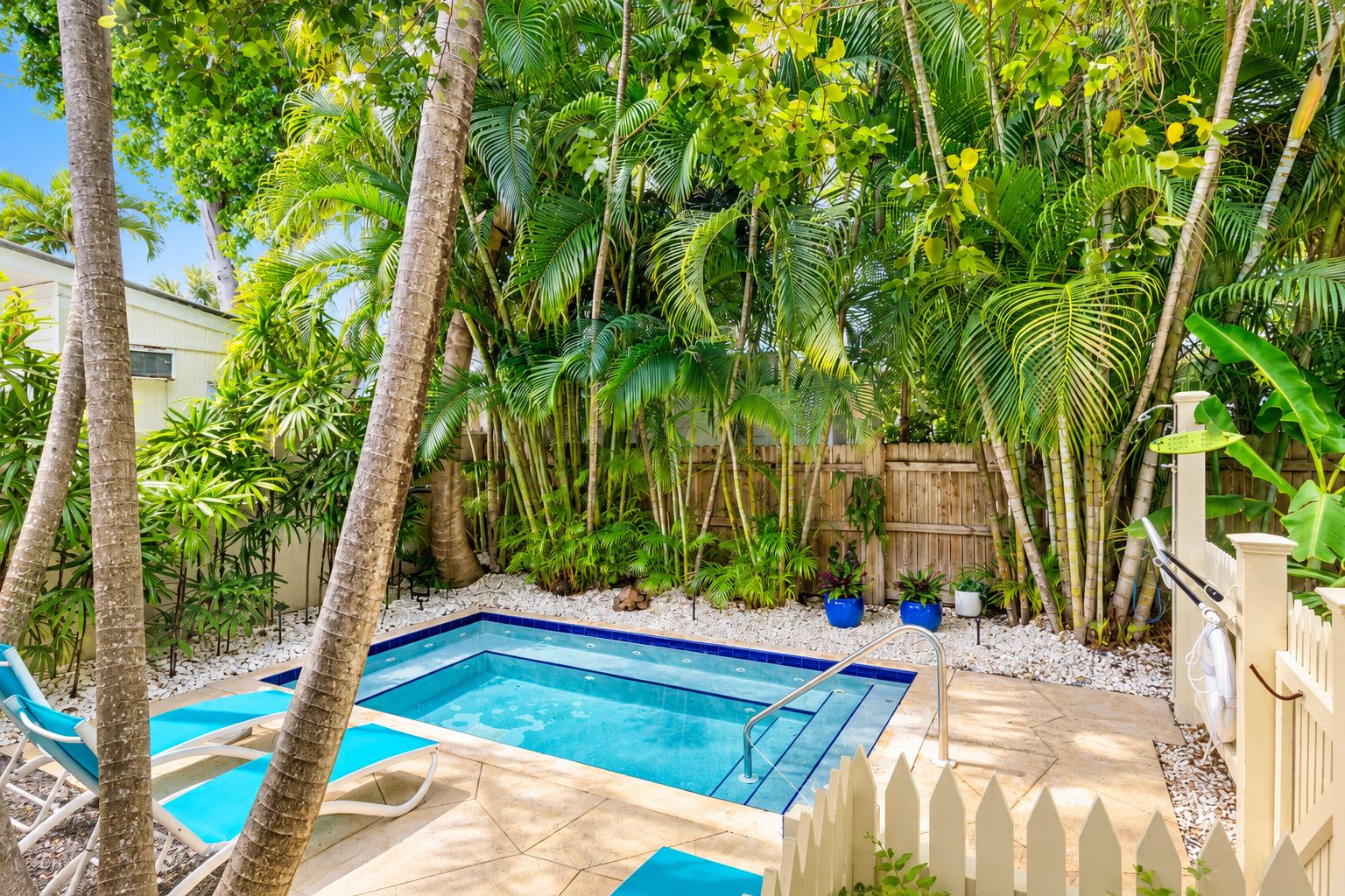 A swimming pool surrounded by tropical vegetation, with a wooden fence and a white picket fence.