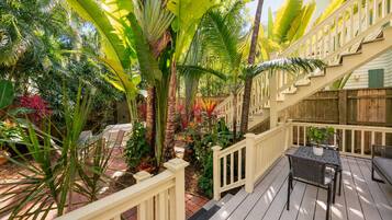 A wooden deck with a railing, surrounded by lush tropical plants and a staircase leading to an upper level.