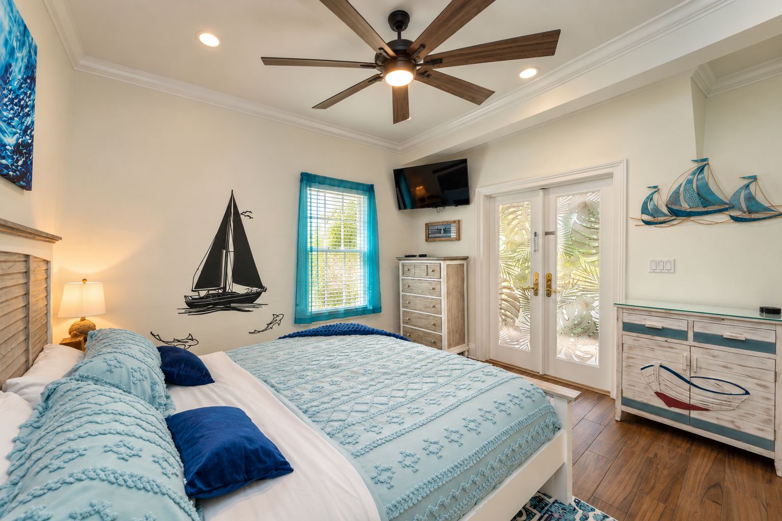 A bedroom with a bed, a ceiling fan, a window with blinds, a TV on the wall, and a cabinet with decorative items.