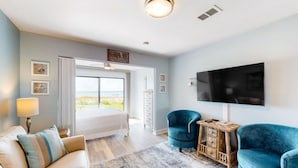 Un dormitorio moderno con una cama, dos sillones azules, una mesita de madera, un televisor y un ventanal con vistas.