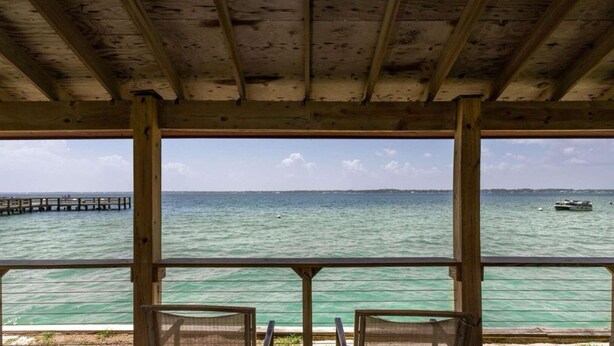 Una terraza de madera con dos sillas que dan a un mar tranquilo y un muelle a lo lejos.