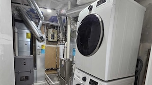 A laundry room with stacked washers and dryers, visible pipes, and a water heater. - Cozy 1BR Suite, Sleeps 3, Private Entry + Parking (Calgary)