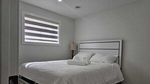 A neatly made bed with white linens and pillows, a bedside table with a lamp, and a window with blinds. - Cozy 1BR Suite, Sleeps 3, Private Entry + Parking (Calgary)