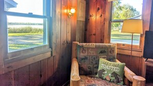 A cozy cabin interior with a wooden chair, a window with a view of a grassy field, and a wall-mounted lamp.
