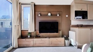 Un salon moderne doté d’une grande fenêtre, d’un téléviseur à écran plat fixé au mur, d’un banc intégré avec espace de rangement et d’un coin cuisine avec des placards et un plan de travail.
