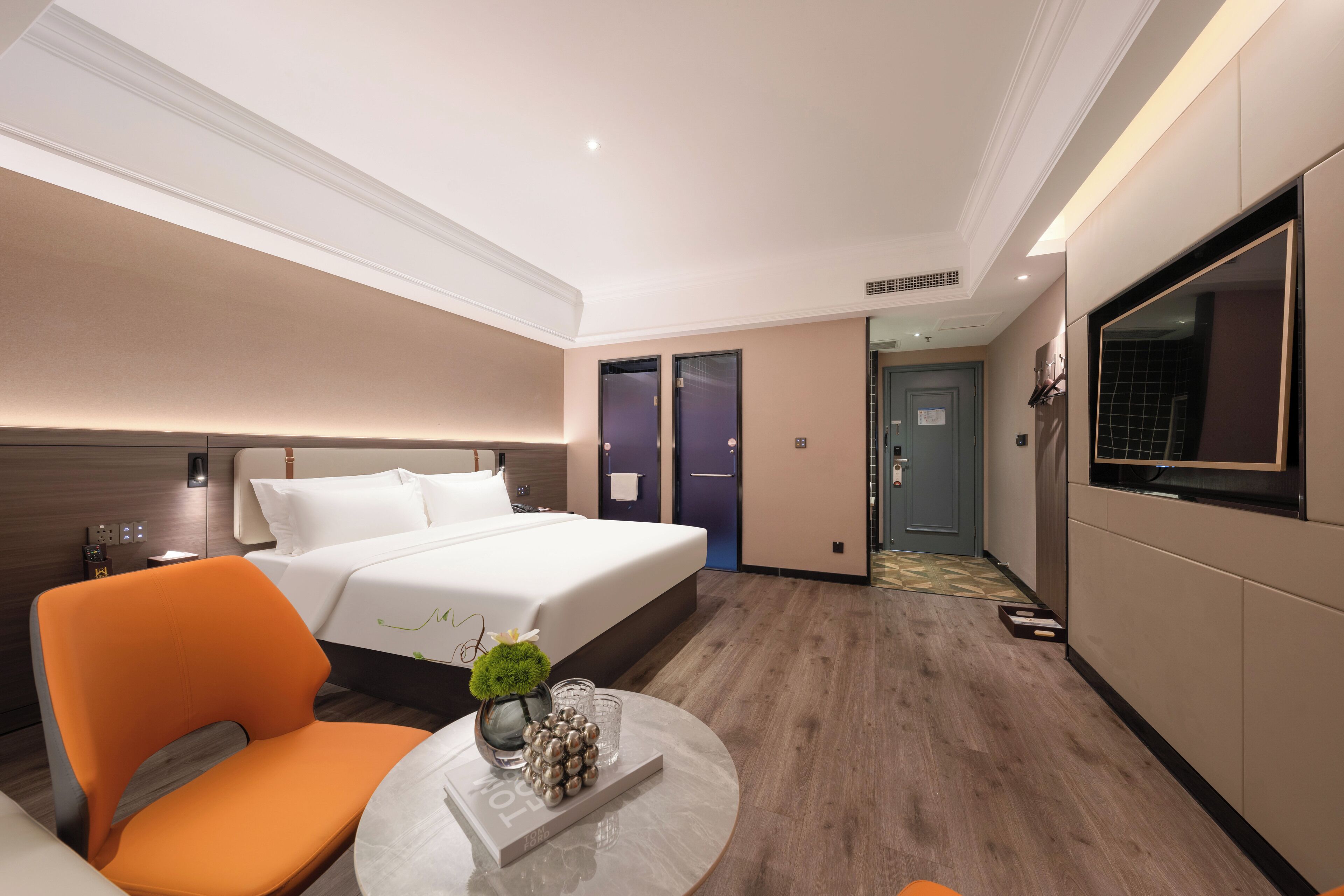 A modern hotel room with a large bed, a seating area with an orange chair, a round coffee table with decorative items, and a TV on the wall.