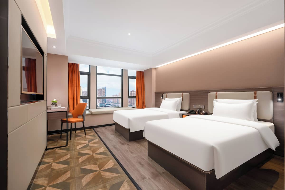 A hotel room with two beds, a desk, a chair, and a large window with a city view.