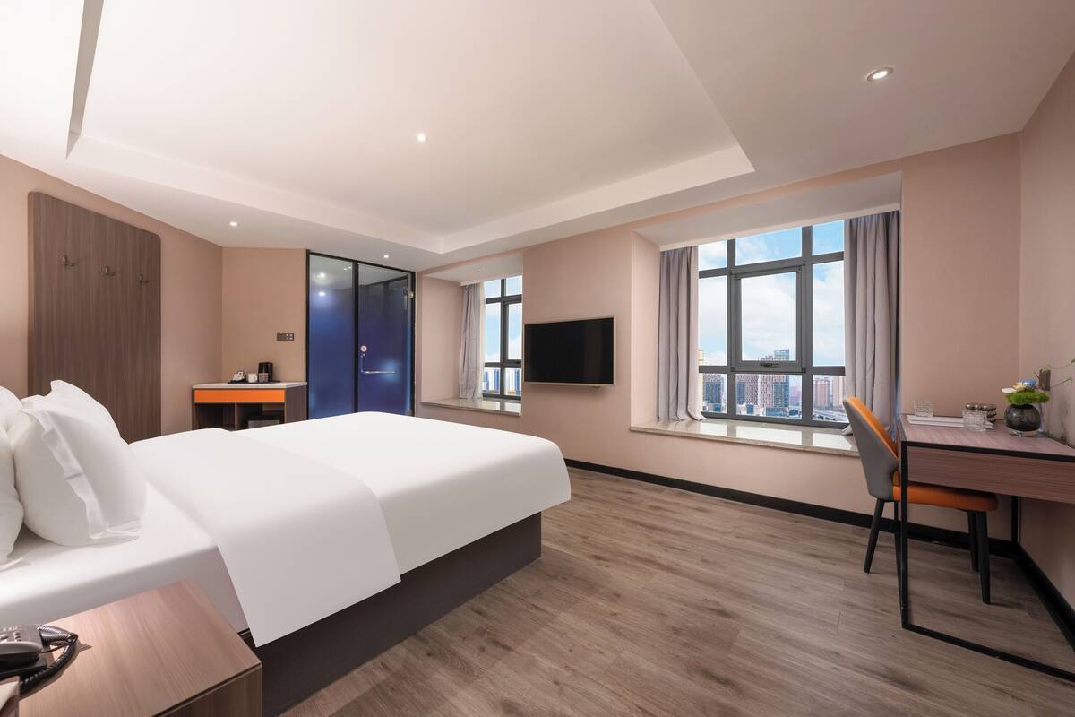 A modern hotel room with a large bed, a desk, a chair, a TV, and a city view.