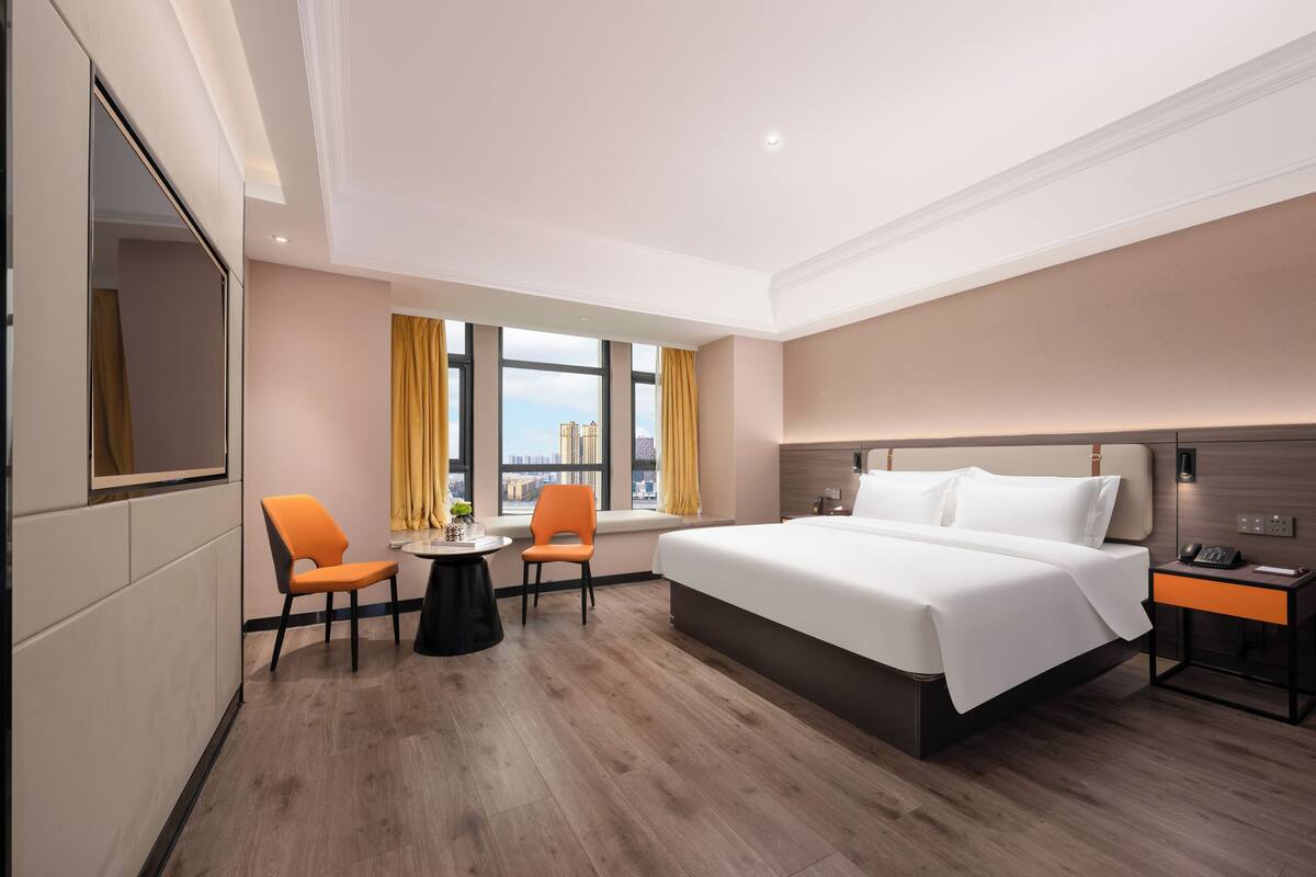 A hotel room with a large bed, two chairs, a small table, a nightstand, and a window with curtains.