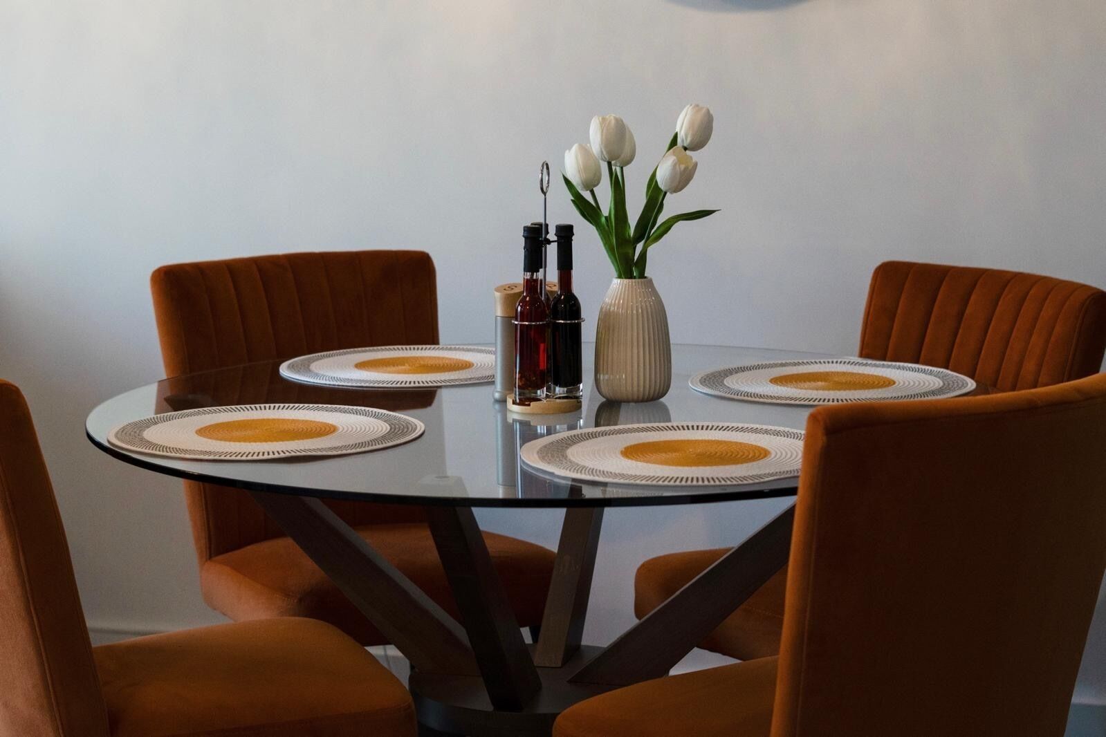 A round glass table with four orange chairs, a vase with white flowers, and two bottles on the table.