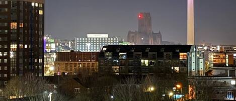 A cityscape at night with a prominent tower with a red light at the top, surrounded by buildings and a parking area.