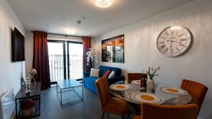 Ein modernes Wohnzimmer mit einem blauen Sofa, orangefarbenen Sesseln, einem runden Esstisch und einer Uhr sowie einem Balkon mit Vorhängen.