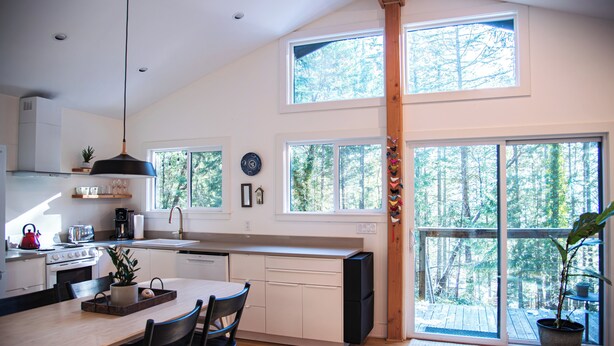 A modern kitchen with a dining area, large windows, and a view of the forest. - Bird House: Fully Renovated Vesuvius Cottage (Salt Spring Island)
