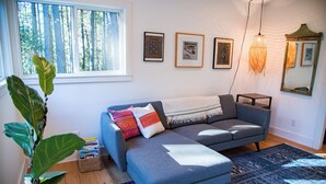 A cozy living room with a gray sectional sofa, a blue rug, and a large window with a view of trees. - Bird House: Fully Renovated Vesuvius Cottage (Salt Spring Island)