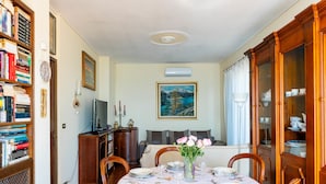 A dining room with a round table set for four, surrounded by wooden chairs. A bookshelf filled with books is to the left, and a painting hangs on the wall above the sofa. - Casa Cycas by Interhome (Ospedaletti)