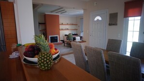Un coin repas avec une table en bois, des chaises et un saladier rempli de fruits. On aperçoit également une cuisine avec des placards et un réfrigérateur au fond.