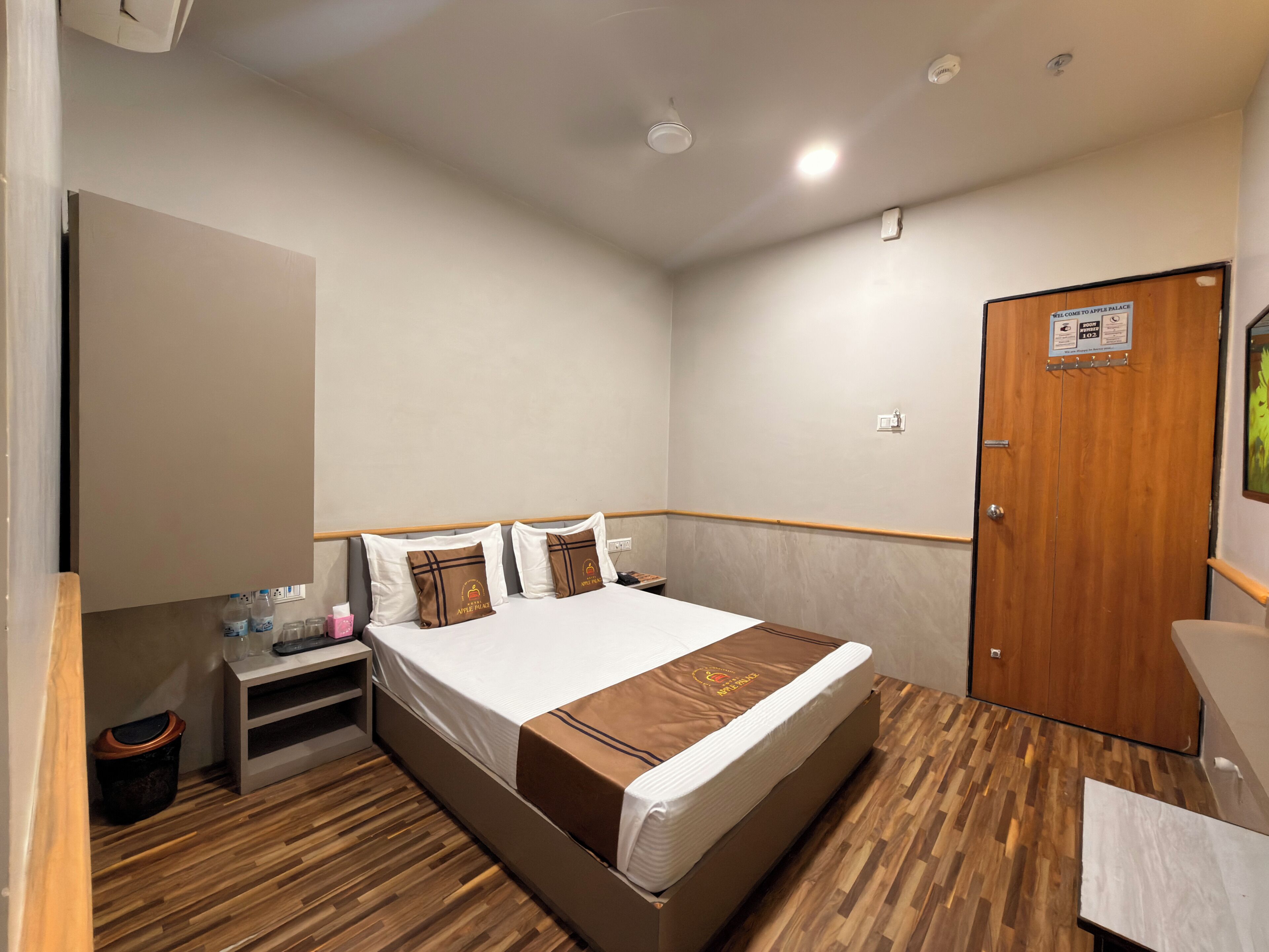 A hotel room with a wooden floor, a bed with white and brown bedding, a nightstand with a bottle and glasses, and a wooden door.