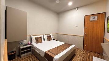 A hotel room with a wooden floor, a bed with white and brown bedding, a nightstand with a bottle and glasses, and a wooden door.