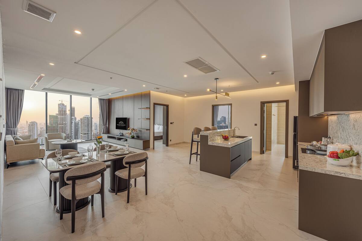 A modern hotel room with a dining area, kitchen, and large windows offering a city view.