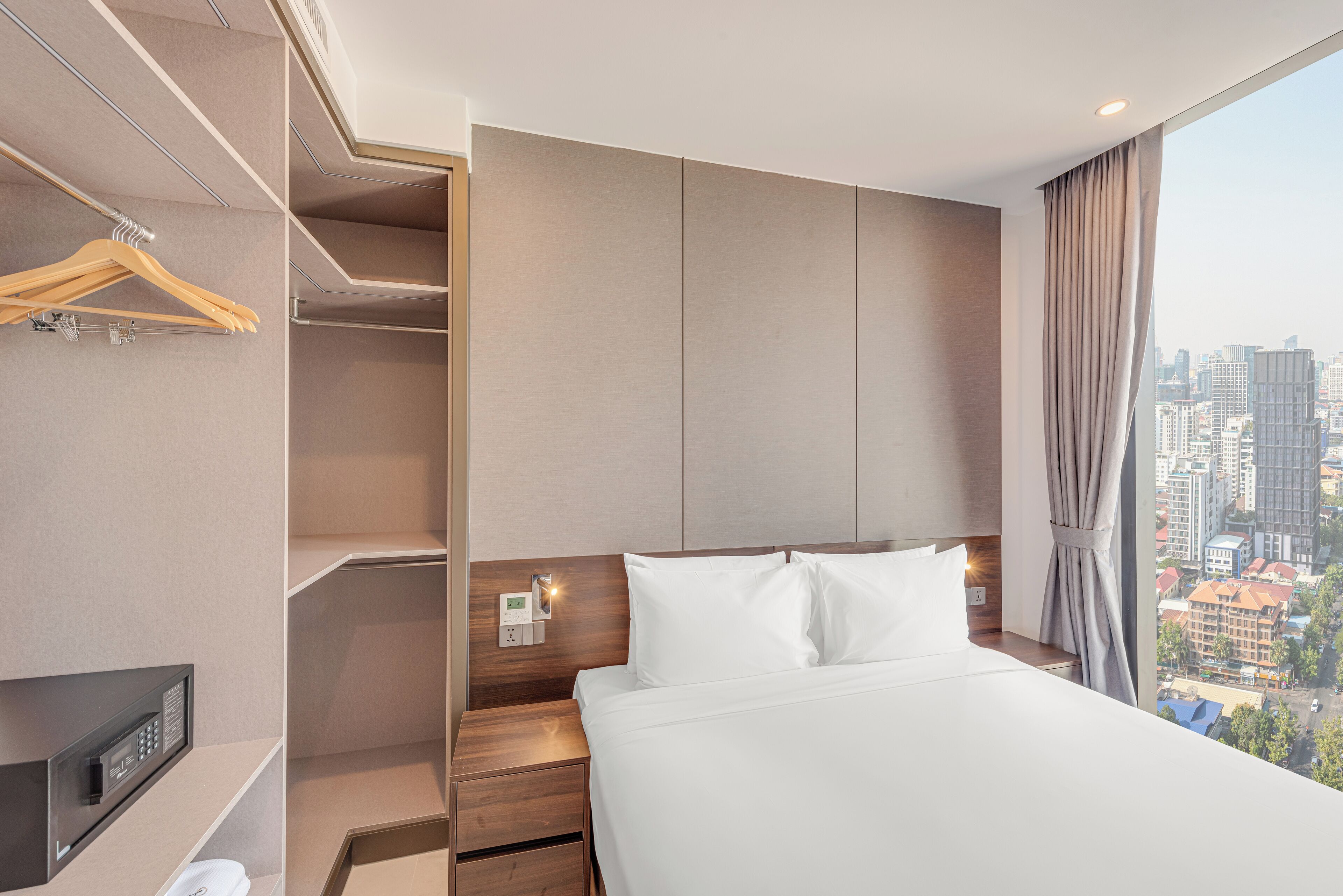 Una habitación de hotel moderna con una cama grande, un vestidor, vistas a la ciudad y un panel de control en la pared.