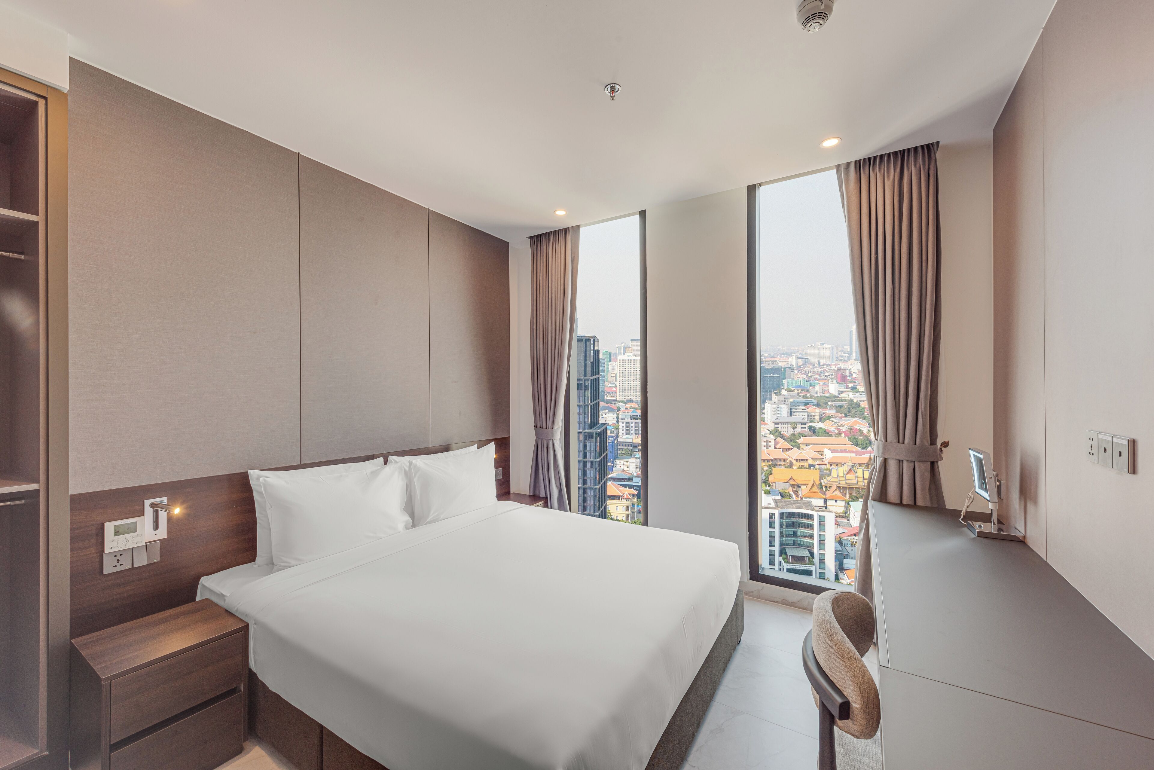Quarto de hotel moderno com cama grande, mesa de cabeceira, escrivaninha e vista para a paisagem urbana através de janelas amplas.