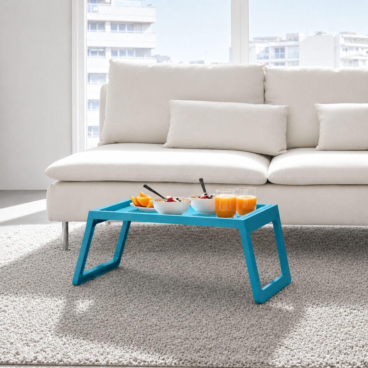 Un salon moderne avec un canapé blanc, une table basse bleue sur laquelle sont disposés des éléments pour le petit-déjeuner, et une vue sur la ville à travers de grandes fenêtres.