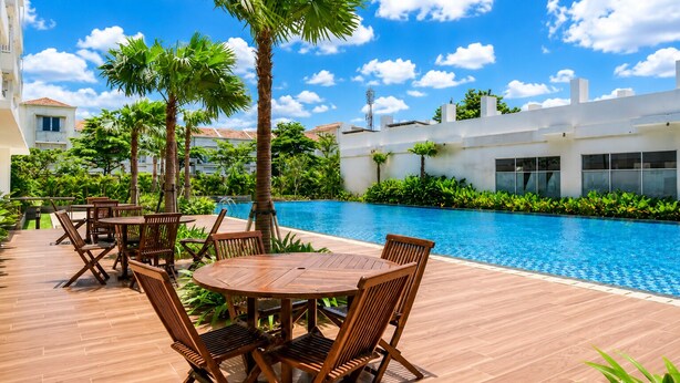 Un espace aménagé au bord de la piscine, avec des tables et des chaises en bois, entouré de palmiers et surplombé d’un ciel bleu azur.