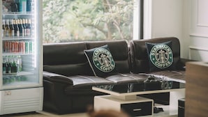 Une pièce avec un canapé en cuir, deux coussins décorés aux couleurs de Starbucks, une machine à café blanche et une fenêtre donnant sur des arbres.