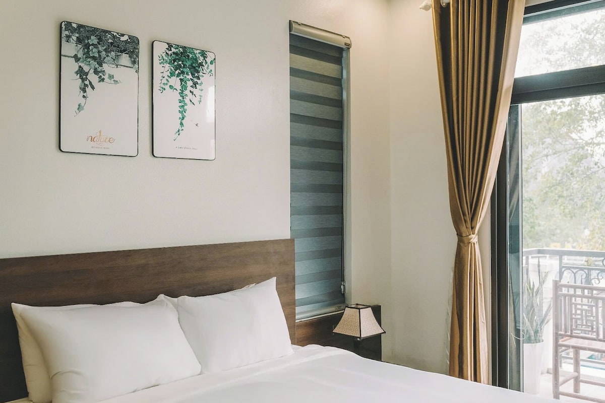 Een net opgemaakt bed met witte lakens, een houten hoofdbord en twee aan de muur bevestigde kunstwerken met bladmotieven.