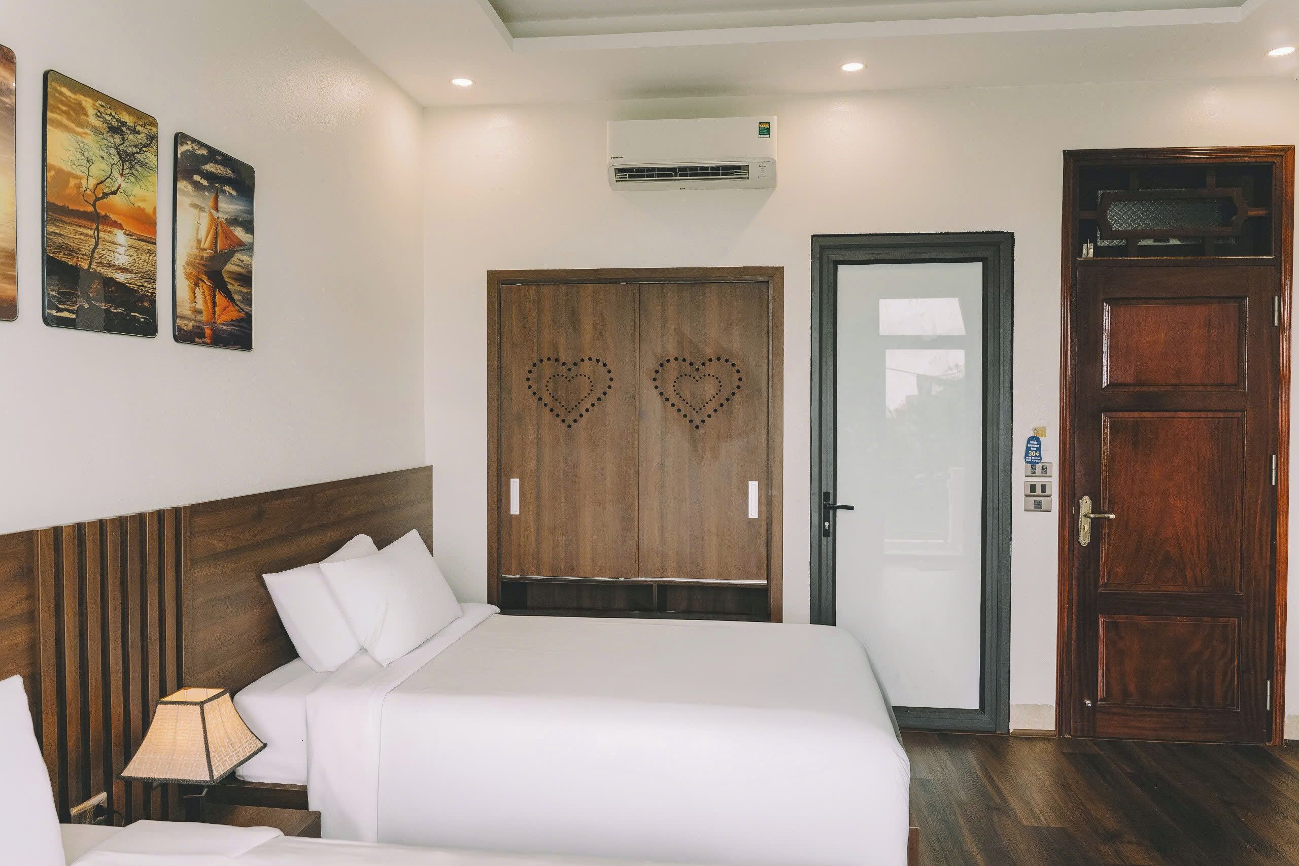 A hotel room with a large bed, a wooden wardrobe with heart-shaped cutouts, a white door, and an air conditioning unit on the wall.