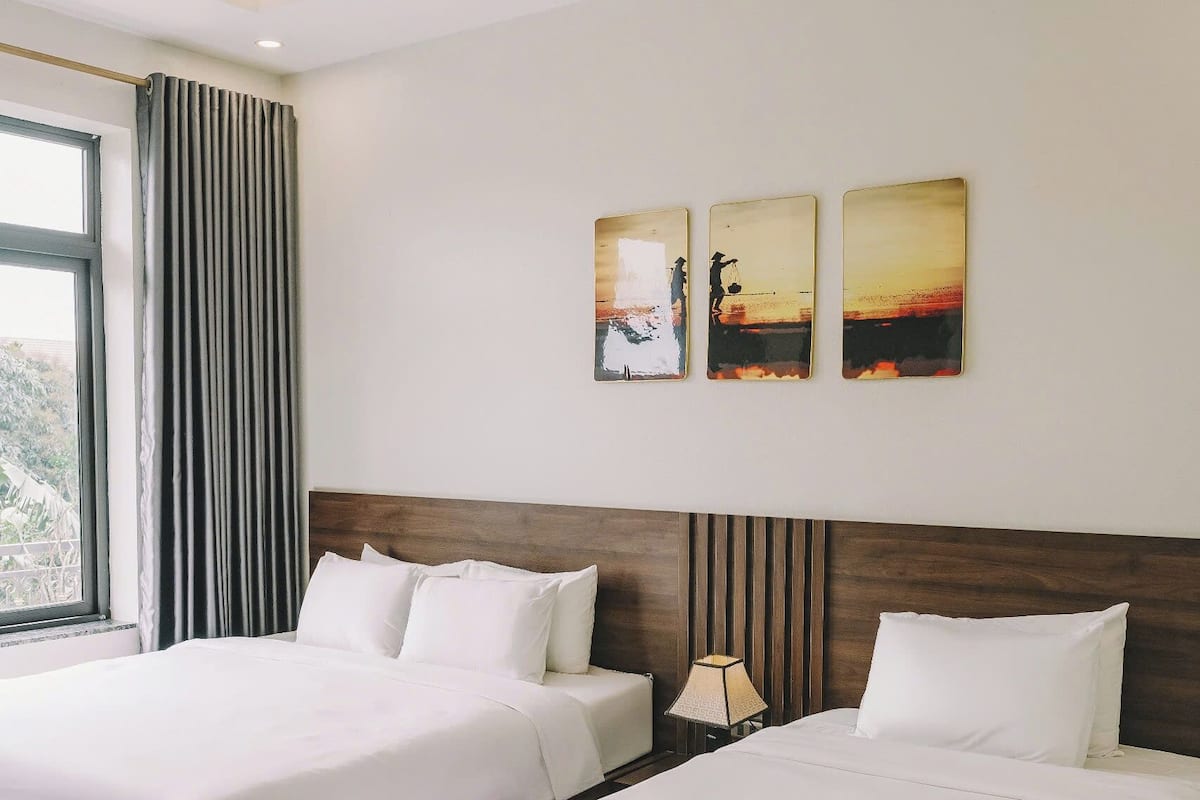 Een hotelkamer met twee bedden, een groot raam met gordijnen, en drie aan de muur bevestigde schilderijen.
