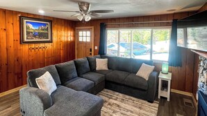 A living room with a sectional sofa, a ceiling fan, wooden walls, a window with a view, and a small table with a lamp. - Yosemite Sky Getaway - Newly Remodeled, 2 King Beds, EV Charger, RV/Boat Parking (Oakhurst)