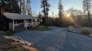 A cabin with a porch, surrounded by a gravel driveway, and a forested area. - Yosemite Sky Getaway - Newly Remodeled, 2 King Beds, EV Charger, RV/Boat Parking (Oakhurst)