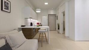 Un salon moderne et minimaliste, avec un canapé, un petit coin cuisine doté d’une table à manger, et un couloir menant à une porte.
