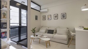 Un salon moderne avec un canapé, une table basse et un téléviseur. Des tableaux encadrés ornent le mur, et un balcon offre une vue agréable.