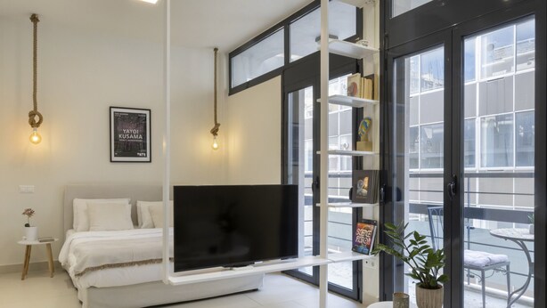 Une chambre moderne avec un lit, un téléviseur fixé au mur blanc, une petite table avec une plante et un balcon avec une chaise.