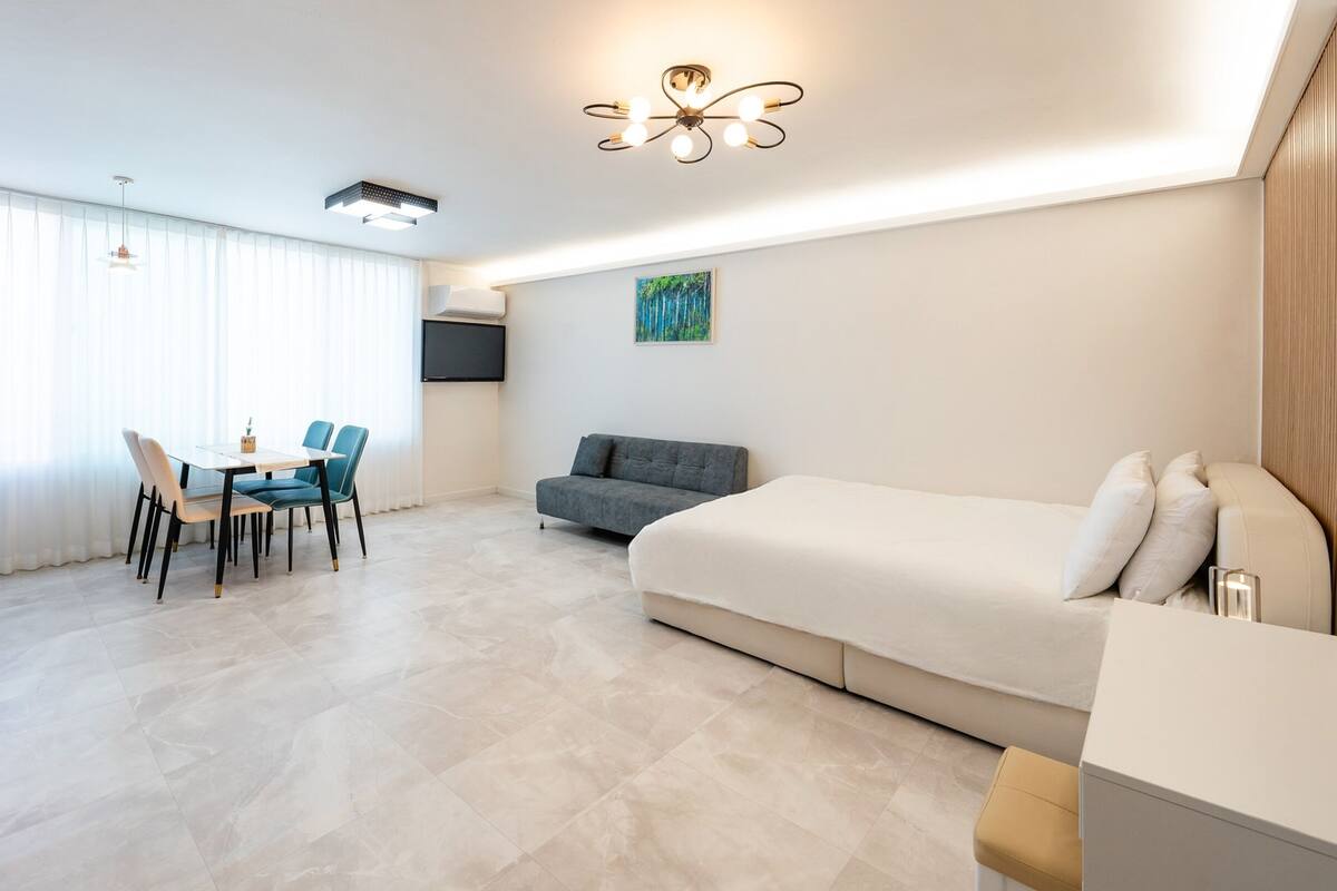 A modern hotel room with a bed, a sofa, a dining table with chairs, a flat-screen TV, and a ceiling light fixture.