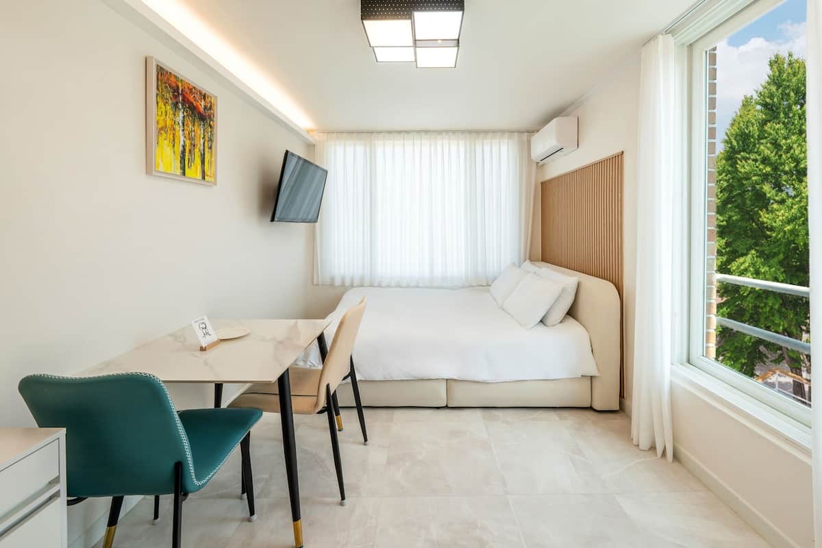 A compact hotel room with a bed, a desk, a chair, a TV, and a window with a view of trees.