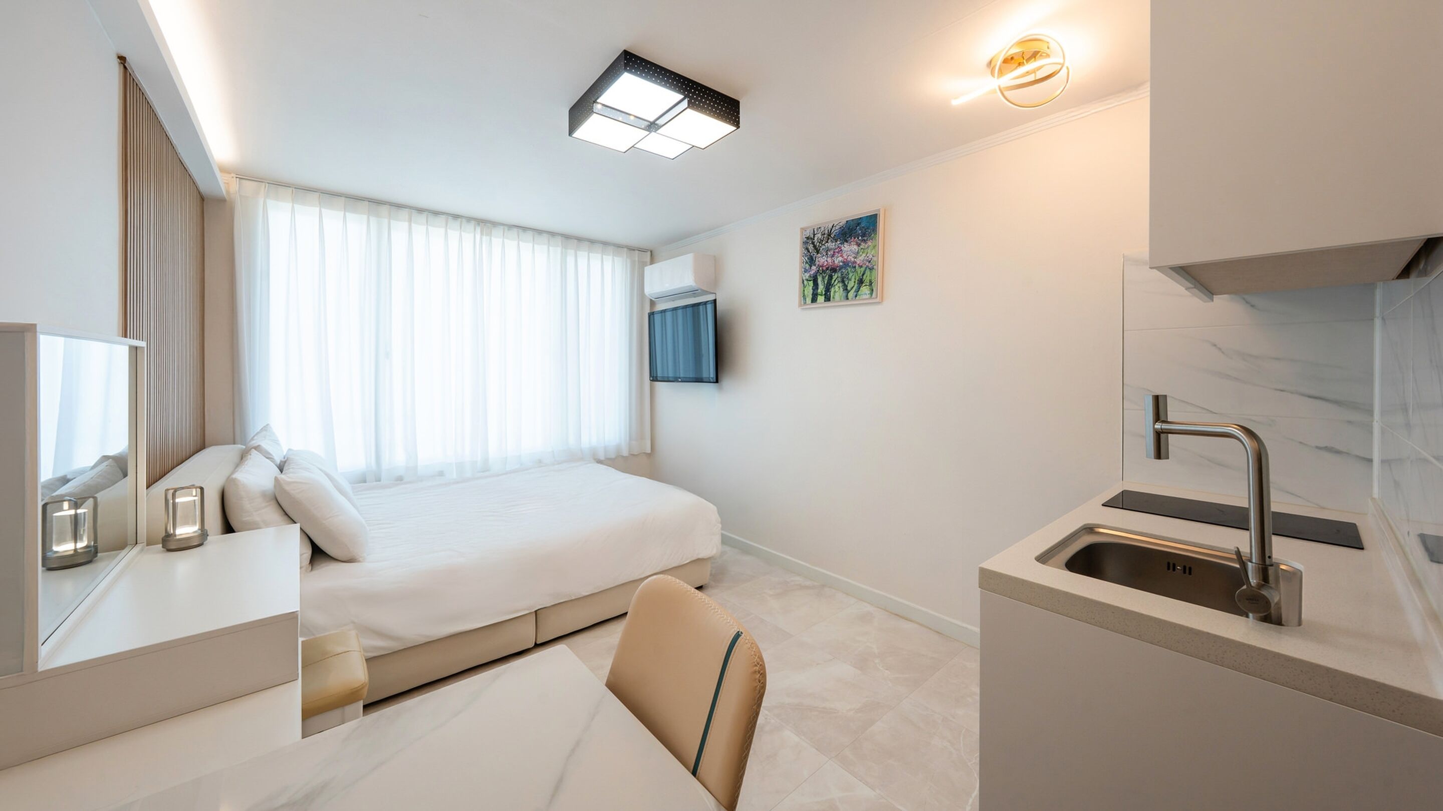 Quarto de hotel moderno com uma cama grande, mesa de cabeceira com luminária, uma cadeira pequena, pia embutida e televisão na parede.