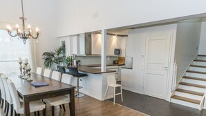 Una cocina moderna con armarios blancos, un comedor con mesa y sillas de madera, y una escalera que conduce a otro nivel.